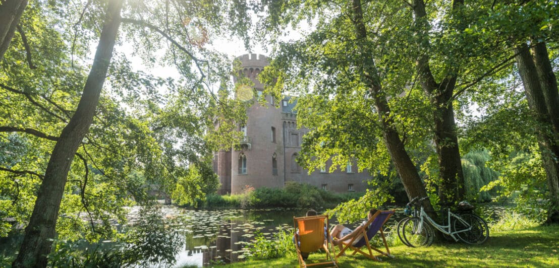Zwei Menschen auf Liegestühlen, die eine Burg in einem See betrachten; an einem Baum lehnen zwei Fahrräder