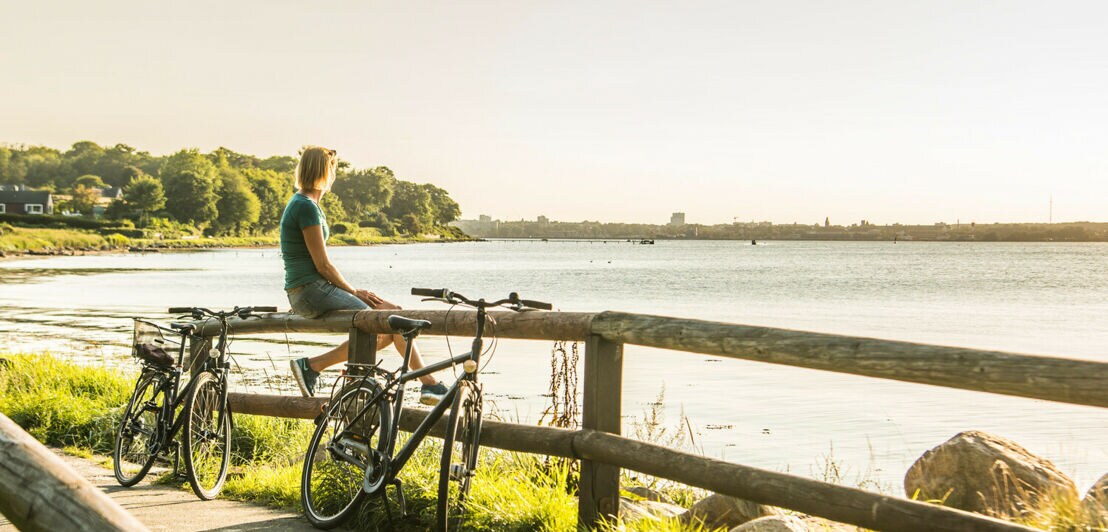 Zwei Fahrräder an einem Holzzaun, auf dem eine Frau sitzt und das Meer betrachtet