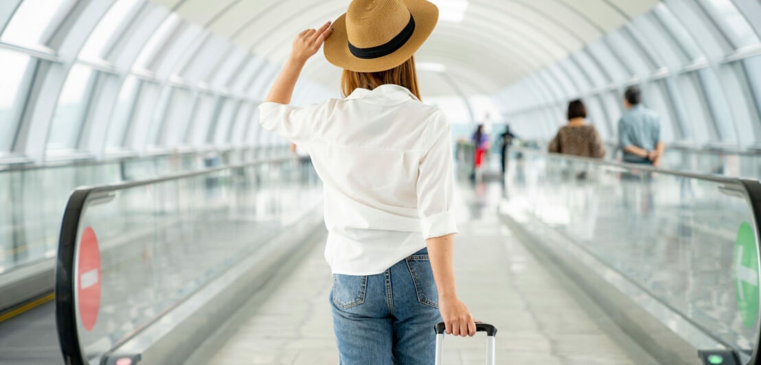 Rückansicht einer reisenden Frau im weißen Hemd mit Strohhut, die einen Trolley im Flughafen hinter sich herzieht