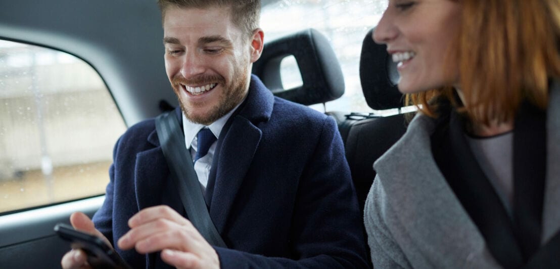 Ein elegant gekleidetes Paar mittleren Alters schaut lachend auf ein Smartphone auf der Rückbank eines Autos.