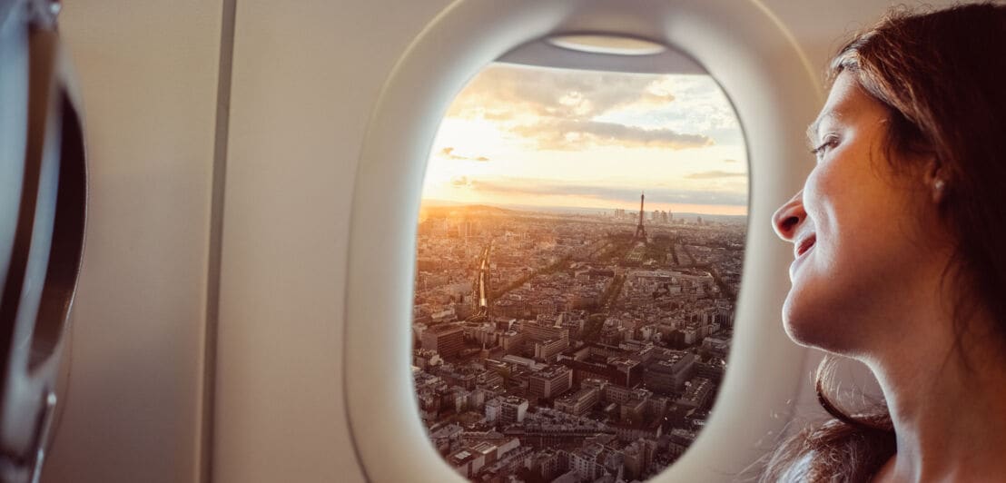 Das Gesicht im Anschnitt einer Frau, die durch ein Flugzeugfenster auf das Stadtpanorama von Paris bei Sonnenuntergang blickt.