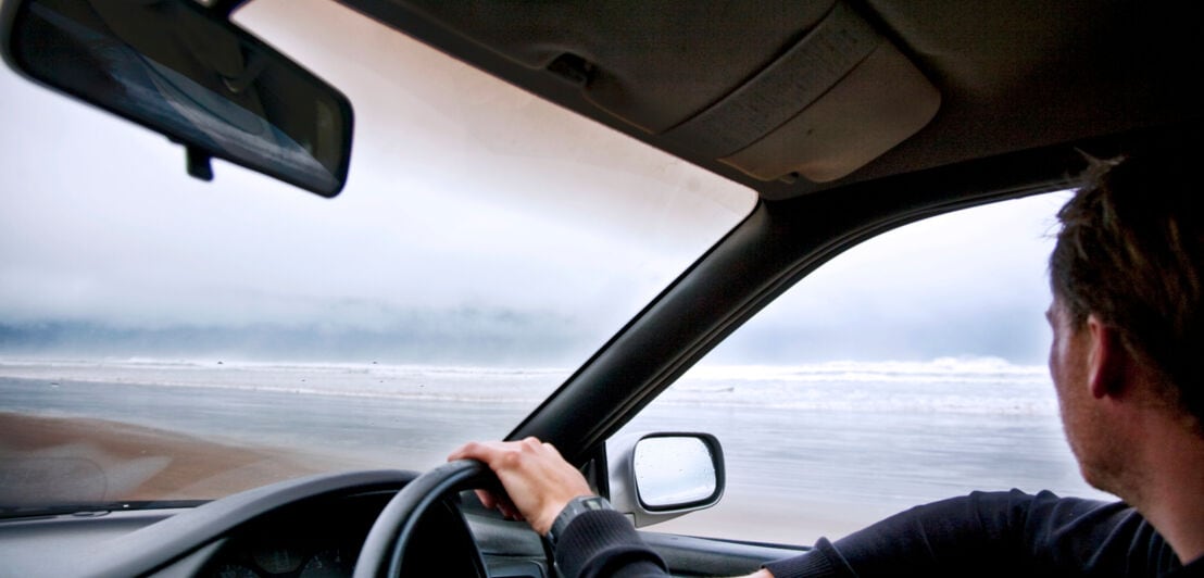Person im Profil am Steuer eines rechtsgelenkten Autos an einem nebeligen Strand.