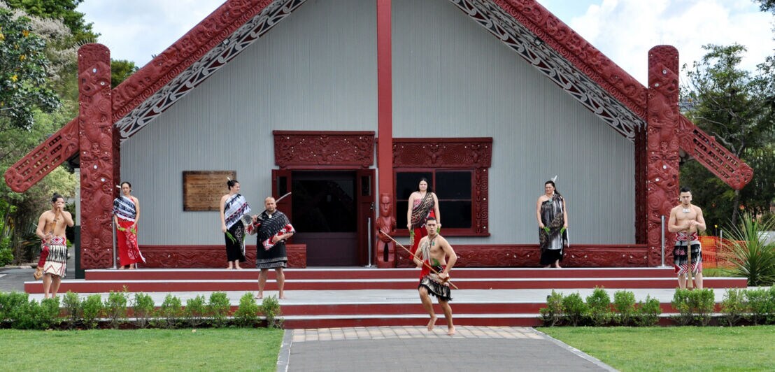 Ein Gruppe Maori bei einer Tanzvorführung vor einem traditionellen Maori Haus.