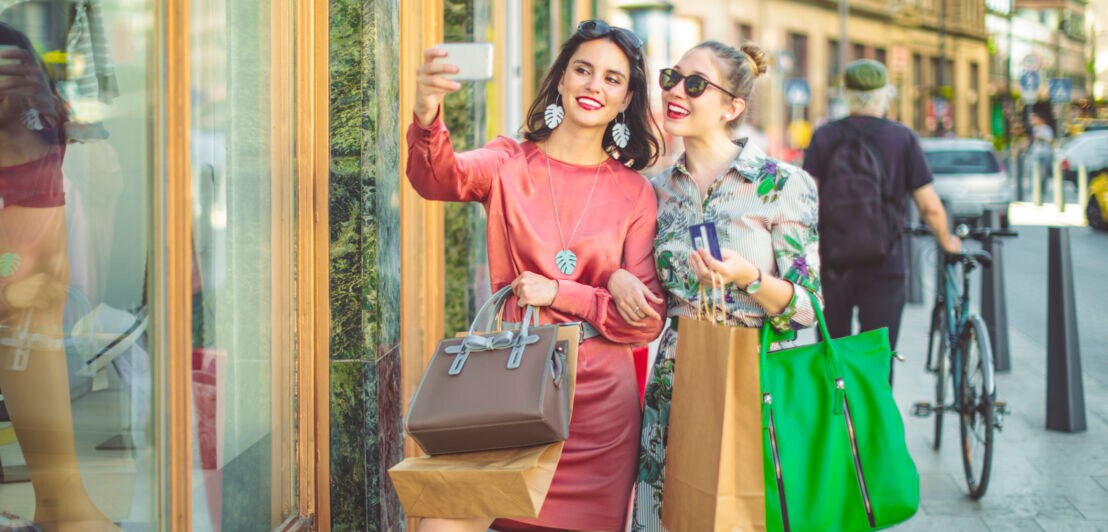 Zwei elegant gekleidete, junge Frauen mit Einkaufstüten machen ein gemeinsames Selfie auf der Straße vor einem Schaufenster.