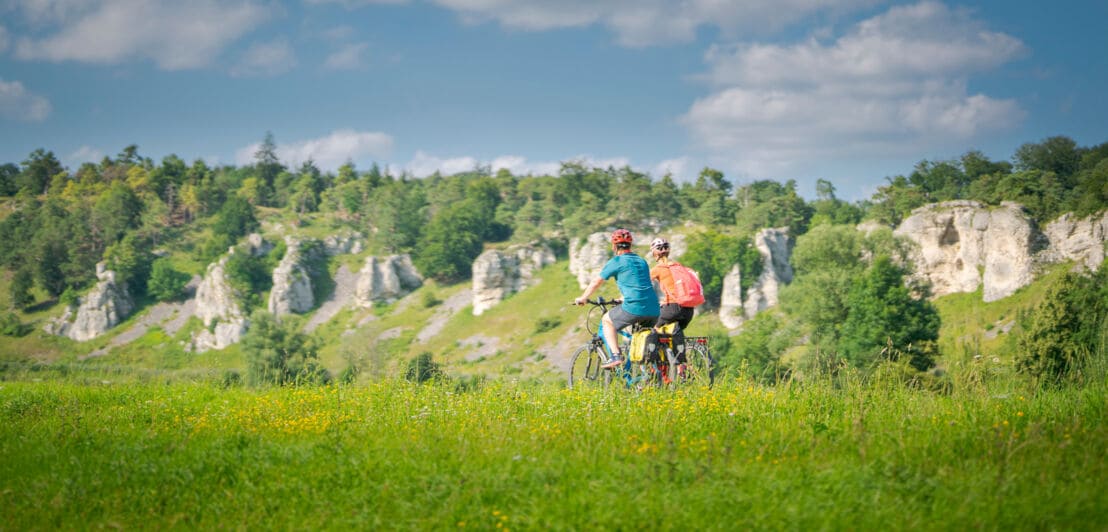 Zwei Personen auf Fahrrädern fahren durch eine Wiesenlandschaft mit kleineren Felsen