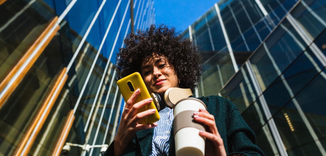 Untersichtaufnahme einer jungen Frau mit Kopfhörern um den Hals, die auf ein gelbes Smartphone in ihrer Hand schaut, während sie in der anderen Hand einen Kaffeebecher hält, im Hintergrund ein modernes Bürogebäude mit Glasfassade