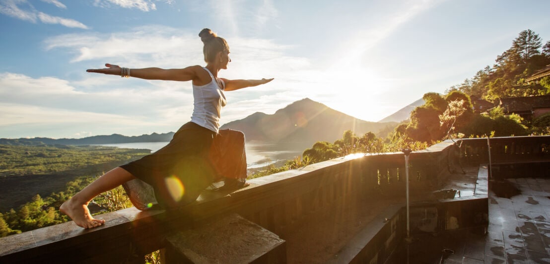 Eine Frau hält eine Yogaposition auf dem Terrassengeländer mit Blick in die weite Landschaft eines Regenwaldes bei Sonnenaufgang