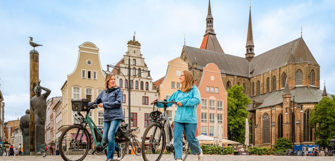 Zwei Frauen schieben nebeneinander ihre E-Bikes, im Hintergrund historische Gebäude in Rostock