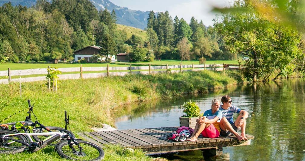 Ein Paar sitzt neben seinen Fahrrädern auf einem Holzsteg an einem idyllischen See vor Bergpanorama im Sonnenschein