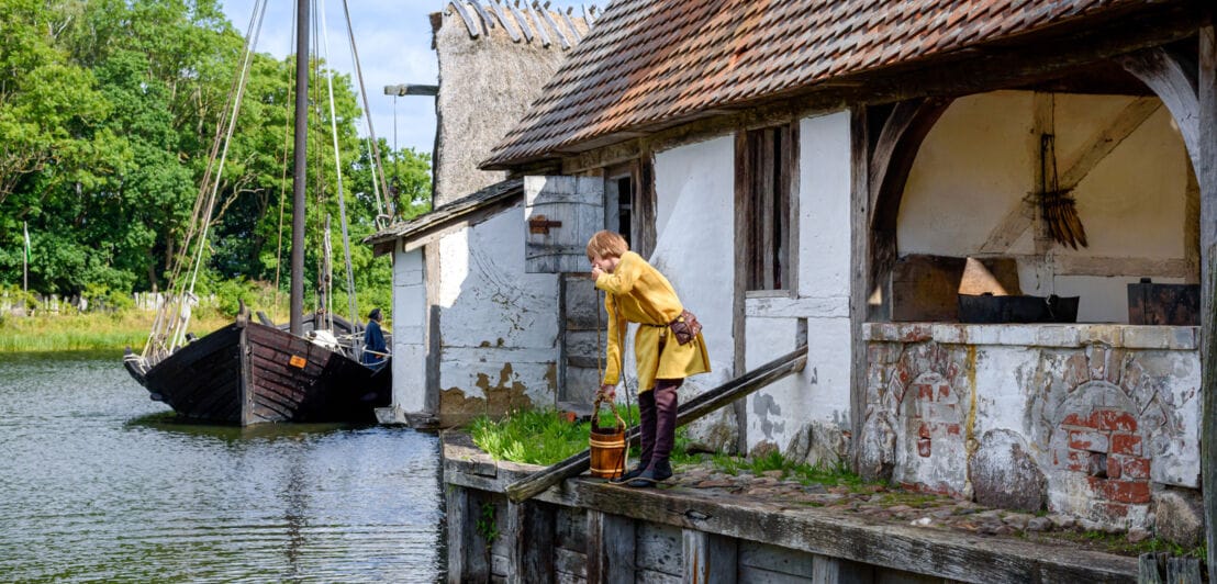 Ein Kind in mittelalterlicher Kleidung seilt einen Holzeimer hinab in einen Fluss in einem Freilichtmuseum
