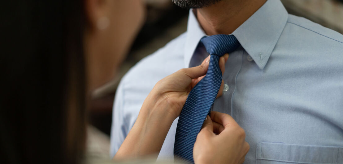 Nahaufnahme von zwei weiblichen Händen, die eine blaue Krawatte am Hals eines Mannes im hellblauen Hemd binden.