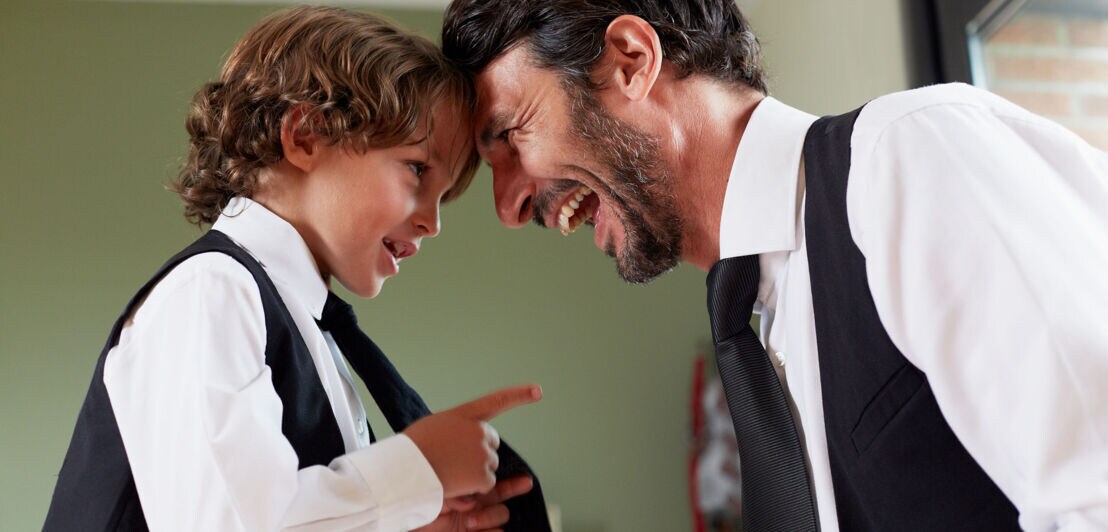 Ein Vater und sein kleiner Sohn im Partnerlook mit weißem Hemd und schwarzer Krawatte stehen sich lachend gegenüber.