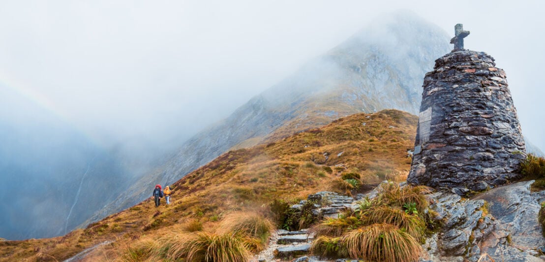 Zwei Wandernde auf dem Milford Track zwischen von Nebel umgebenen Felsen und Graslandschaft