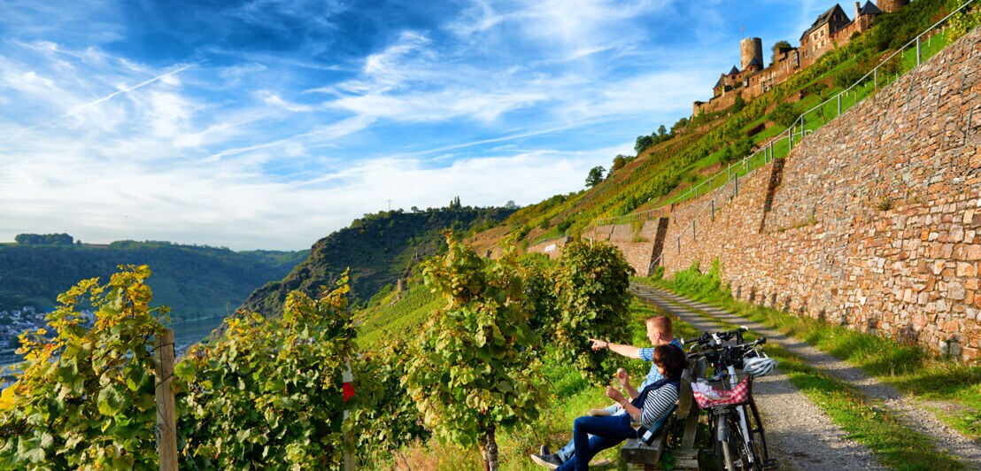 Zwei Personen sitzen auf einer Bank und blicken über Weinreben. Hinter ihnen stehen ihre Fahrräder, auf dem Weinberg ist die Burg Thurant zu erkennen