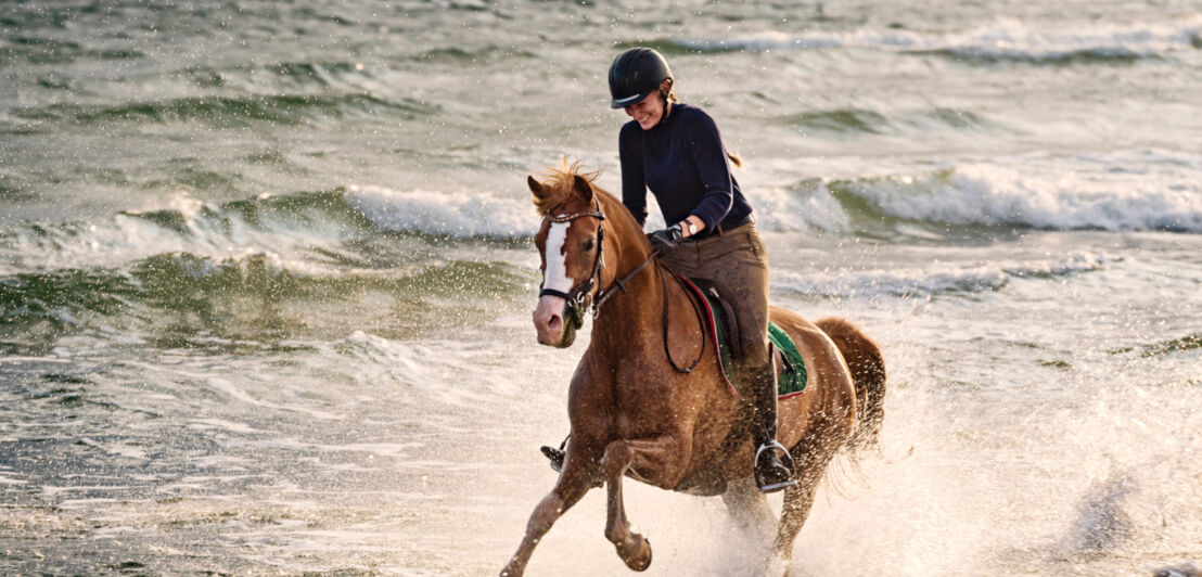 Eine Reiterin galoppiert mit ihrem braunen Pferd durchs Meerwasser