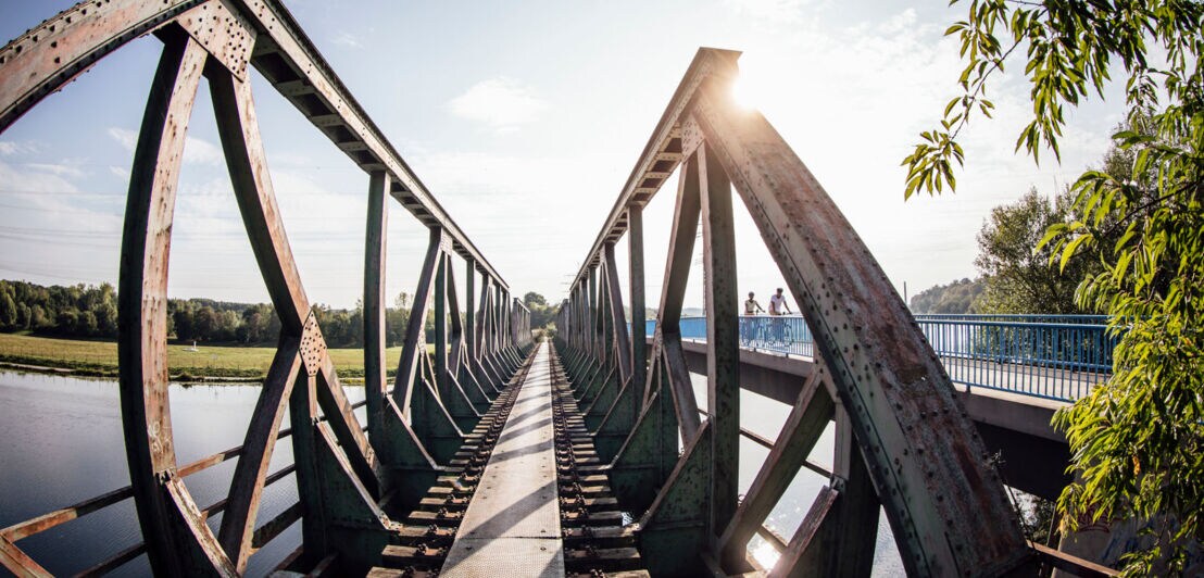 Zwei Radfahrer fahren auf einer Brücke neben einer alten Eisenbahnbrücke über einen Fluss