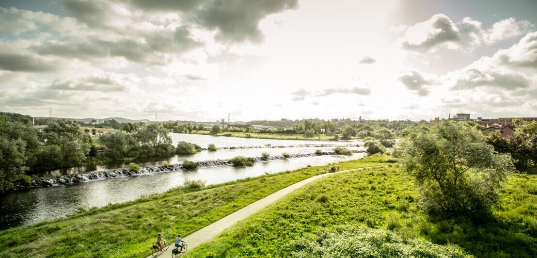 Zwei Radfahrer fahren auf einem Radweg entlang eines Flusses durch eine grüne Auenlandschaft