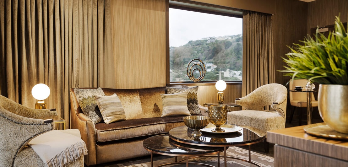 Elegantes Wohnzimmer in Beigetönen mit goldfarbenen Accessoires und Fensterblick in eine bewaldete Hügellandschaft.