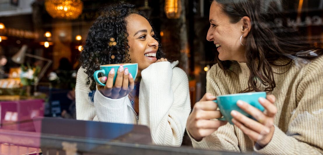 Zwei Frauen mit Kaffeetassen in den Händen sitzen am Fenster eines Cafés und schauen sich fröhlich an.