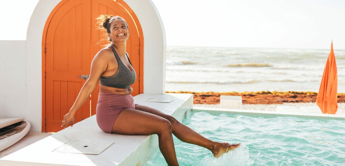 Eine Frau in Sportkleidung sitzt am Rande eines Pools vor Meereskulisse und plantscht gut gelaunt mit den Füßen im Wasser.