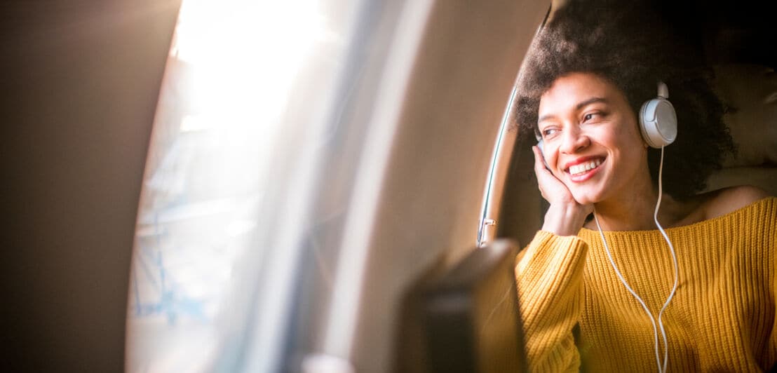 Eine Frau mit gelbem Pullover und weißen Kopfhörern sitzt im Flugzeug und schaut lächelnd aus dem Fenster