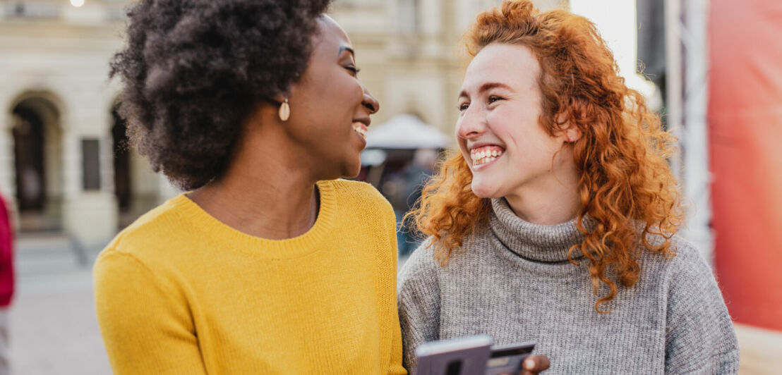 Zwei junge Frauen schauen sich lachend auf der Straße an, eine Frau hält dabei ein Smartphone und eine Kreditkarte in den Händen