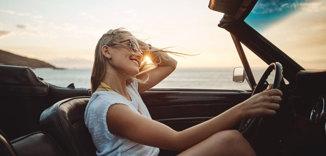 Eine junge Frau mit Sonnenbrille am Steuer eines Cabriolets fährt mit wehenden Haaren eine Küstenstraße bei Sonnenuntergang entlang
