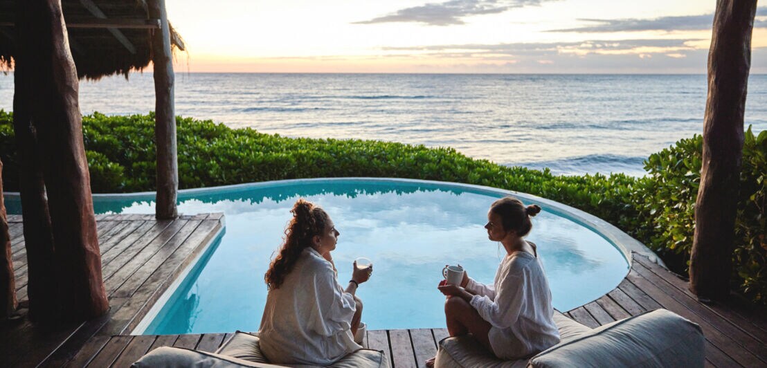 Zwei junge Frauen in weißen Bademänteln sitzen mit Getränken auf Sitzkissen an einem Pool am Meer