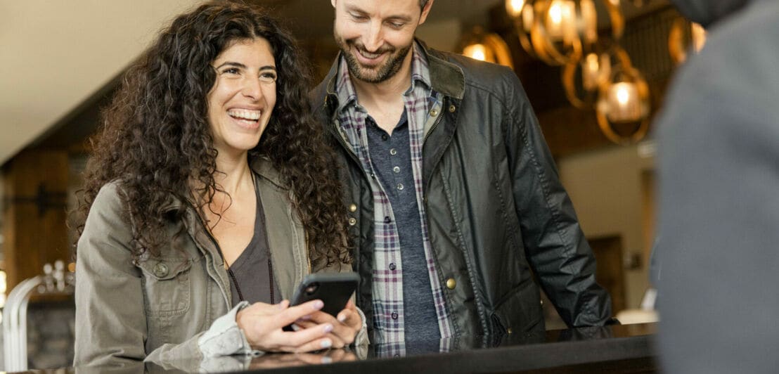 Ein Mann und eine Frau mit Smartphone stehen lächelnd an einem Tresen in einem Foyer
