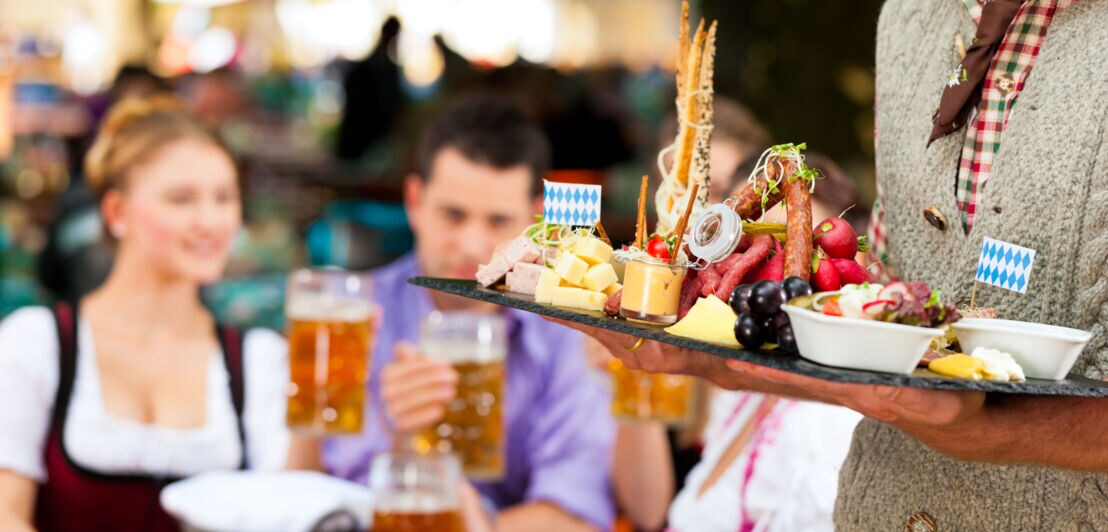 Nahaufnahme einer Vesperplatte in den Händen einer Person in bayerischer Tracht, im Hintergrund eine Personengruppe an einem Tisch mit Bierhumpen in einem Biergarten