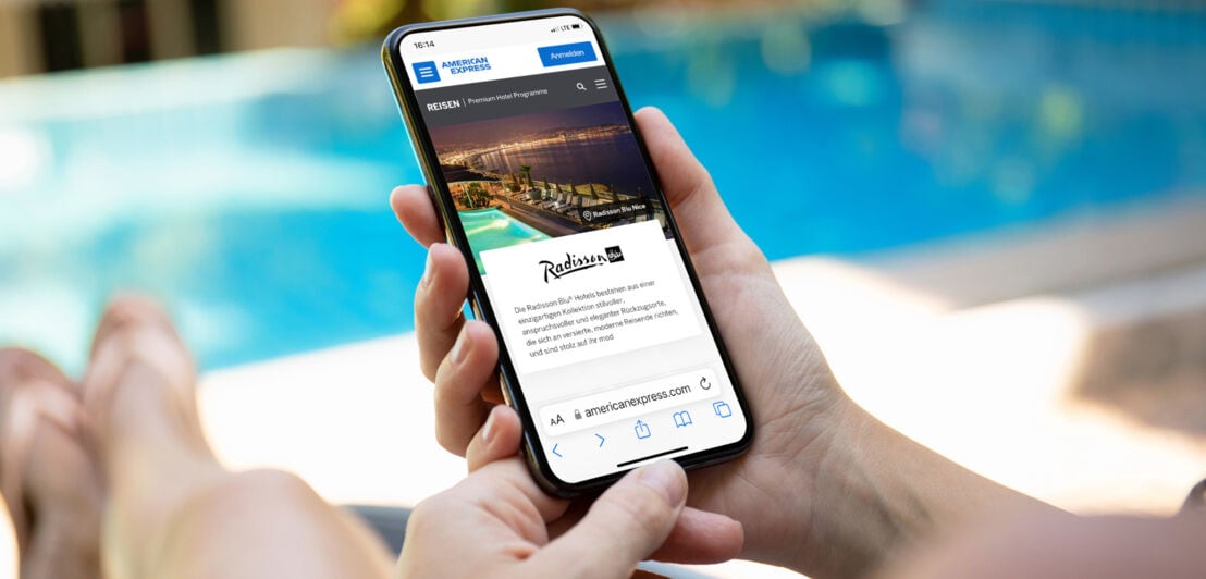 Eine Person auf einer Poolliege hält ein Smartphone mit dem American Express Online-Reiseportal auf dem Display in der Hand
