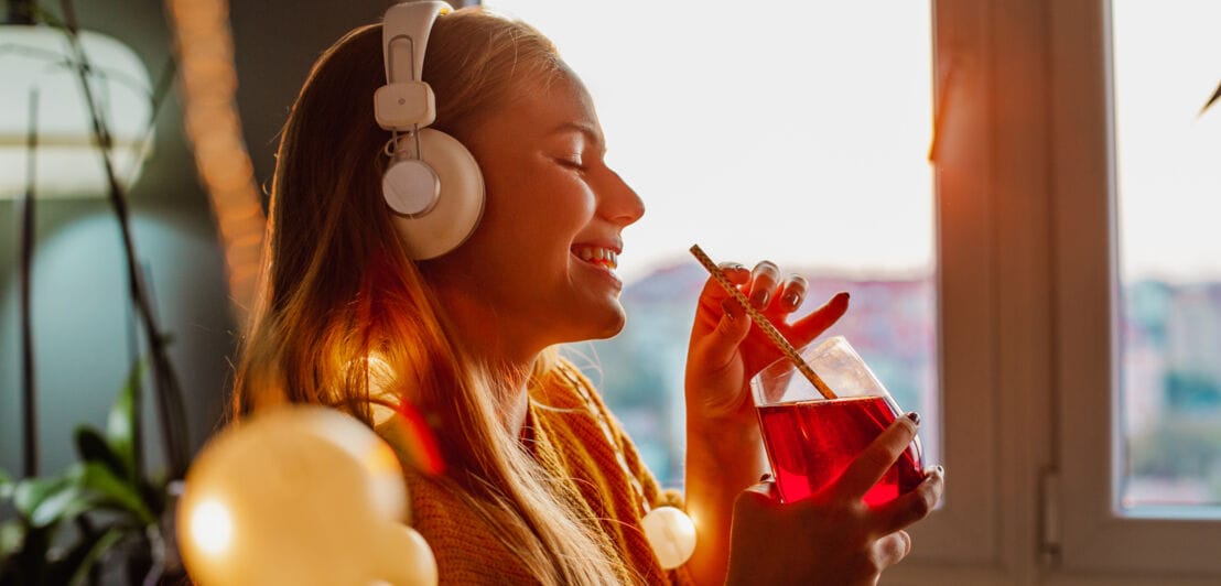 Eine junge, lächelnde Frau mit weißen Kopfhörern genießt ein rotes Getränk mit Strohhalm am Fenster einer Wohnung.