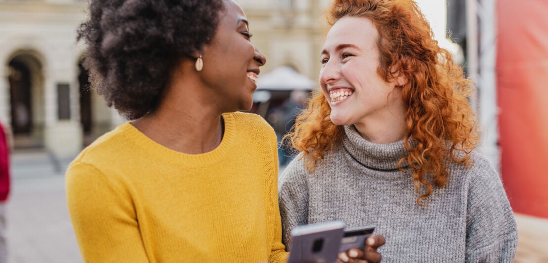 Zwei lachende junge Frauen mit einem Smartphone und einer Kreditkarte in einer Einkaufsstraße.