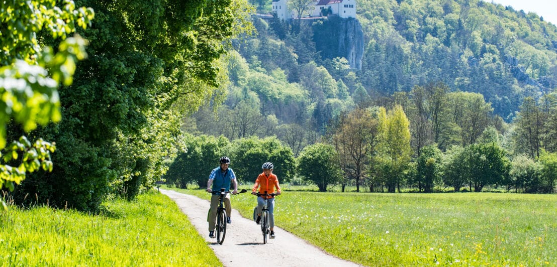 Zwei Personen auf Fahrrädern fahren auf einem Radweg durch ein grünes Tal.