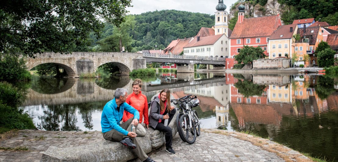 Drei Personen mit Fahrrädern sitzen auf einem Stein am Ufer eines Flusses vor dem Panorama einer mittelalterlichen Kleinstadt.