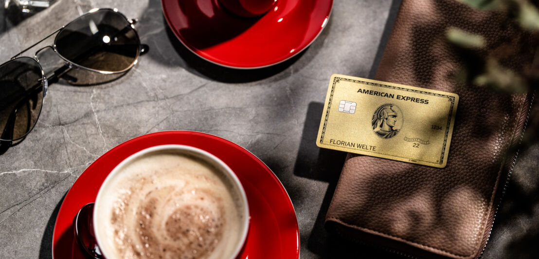 Eine goldene American Express Kreditkarte liegt auf einer Geldbörse auf einem Tisch mit zwei roten Kaffeetassen