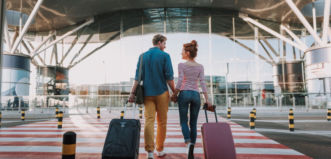 Rückansicht eines händchenhaltenden Paares mit Koffern an einem Flughafenterminal.