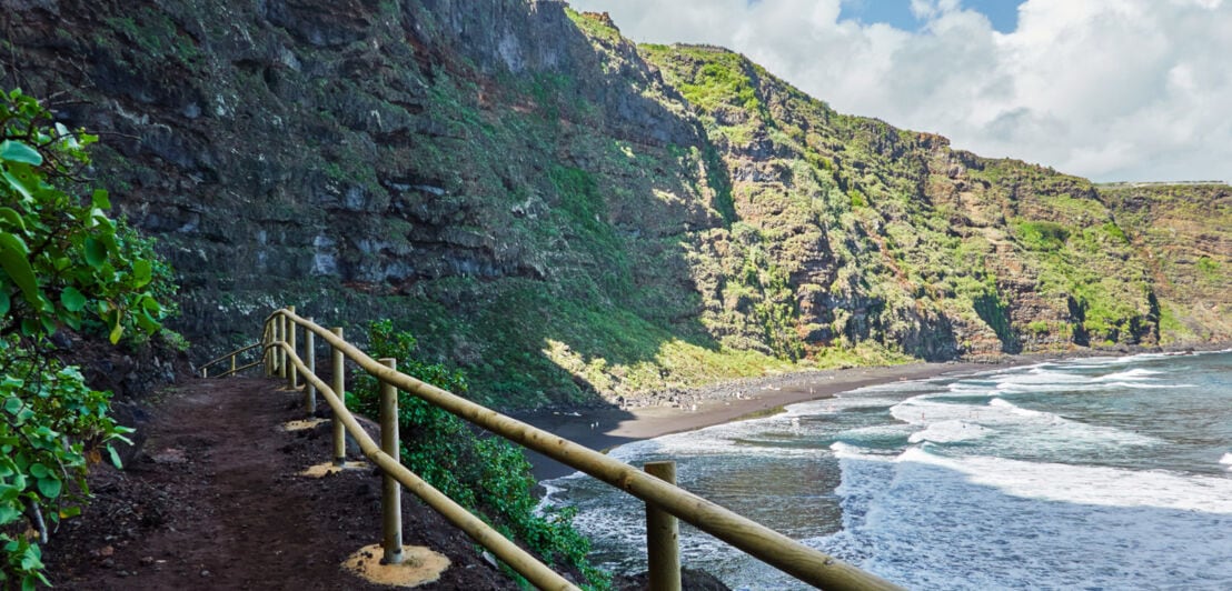 Ein schmaler Wanderweg, der entlang einer grün bewucherten Felsklippe am Meer zu einem kleinen Sandstrand führt