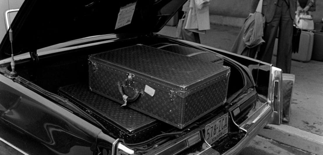 Mehrere, große Lederkoffer mit Monogramm-Muster von Louis Vuitton im Kofferraum eines Cadillacs, Schwarzweißfoto.