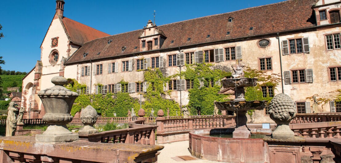 Ein historisches Gebäude, bewachsen mit Grün, davor ein Abteigarten mit steinernen Skulpturen