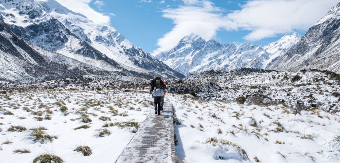 Eine Person in Wandermontur geht einen Weg zwischen schneebedeckten Bergen entlang