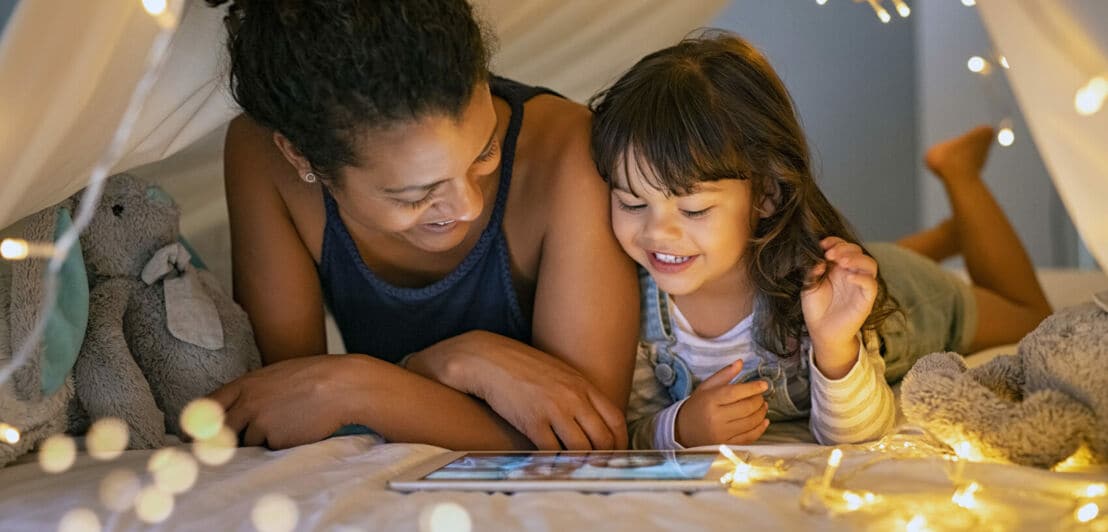 Eine Frau und ein kleines Mädchen liegen in einem Spielzelt mit Lichterketten und schauen gemeinsam einen Film auf einem Tablet an.