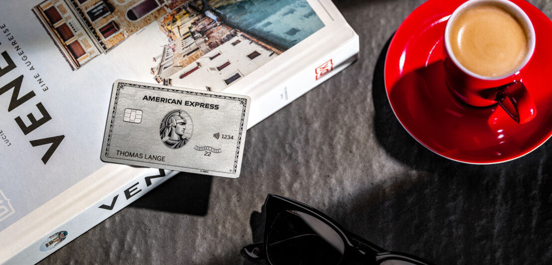 Eine silberne Kreditkarte von American Express liegt auf einem Reisebuch neben einer Espressotasse und einer Sonnenbrille