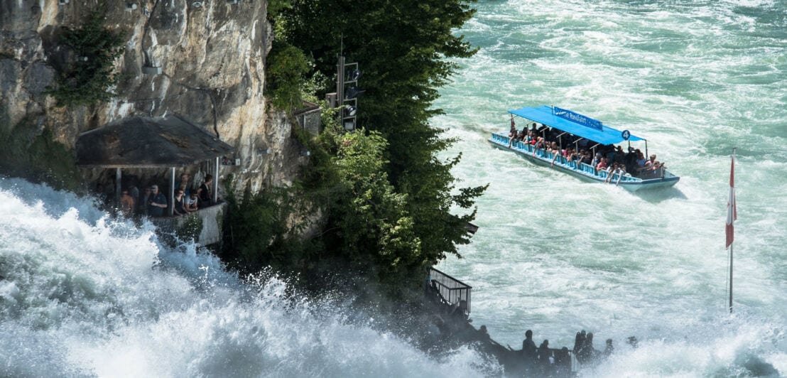 Personen in einem Ausflugsboot und Personen auf einer Aussichtsplattform an einem Wasserfall