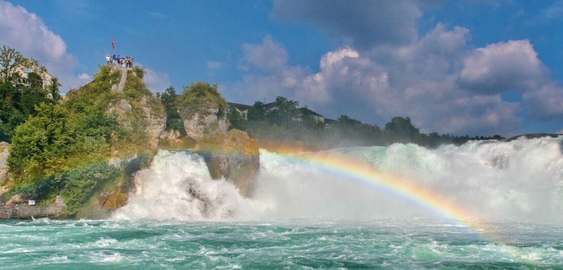 Personen auf einer Aussichtsplattform auf einem Felsen oberhalb eines Wasserfalls, vor dem sich ein Regenbogen gebildet hat