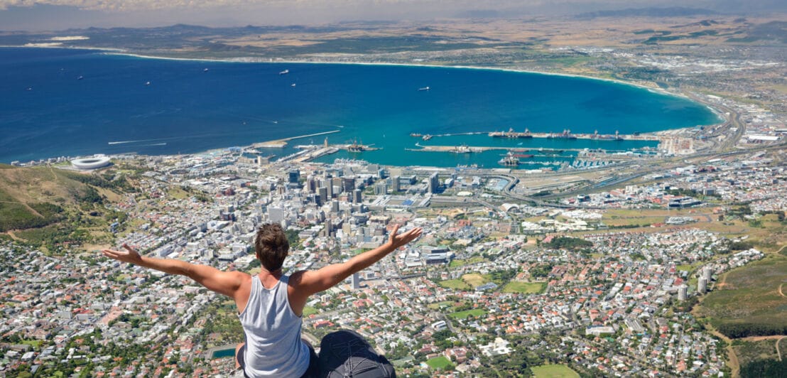 Eine männliche Person sitzt mit dem Rücken zur Kamera auf einem Fels, breitet die Arme aus und blickt auf Kapstadt