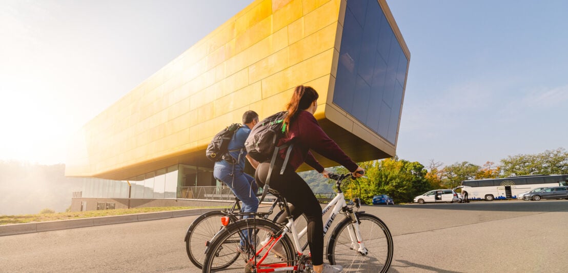 Rückansicht zweier Personen auf Fahrrädern vor einem freistehenden, futuristischen Museumsgebäude