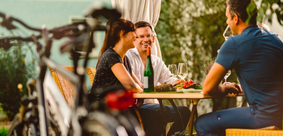 Drei Personen an einem Tisch bei einer Weinverkostung, im Vordergrund geparkte Fahrräder