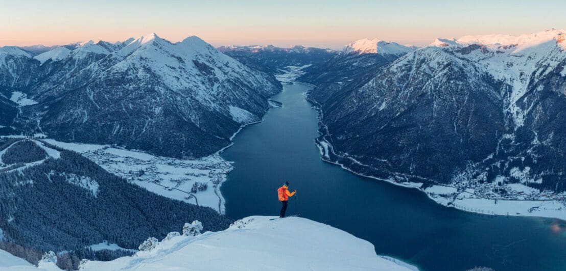 Eine Person steht auf einer schneebedeckten Fläche auf einem Berg, im Hintergrund liegt ein langgezogener See zwischen zwei Bergketten
