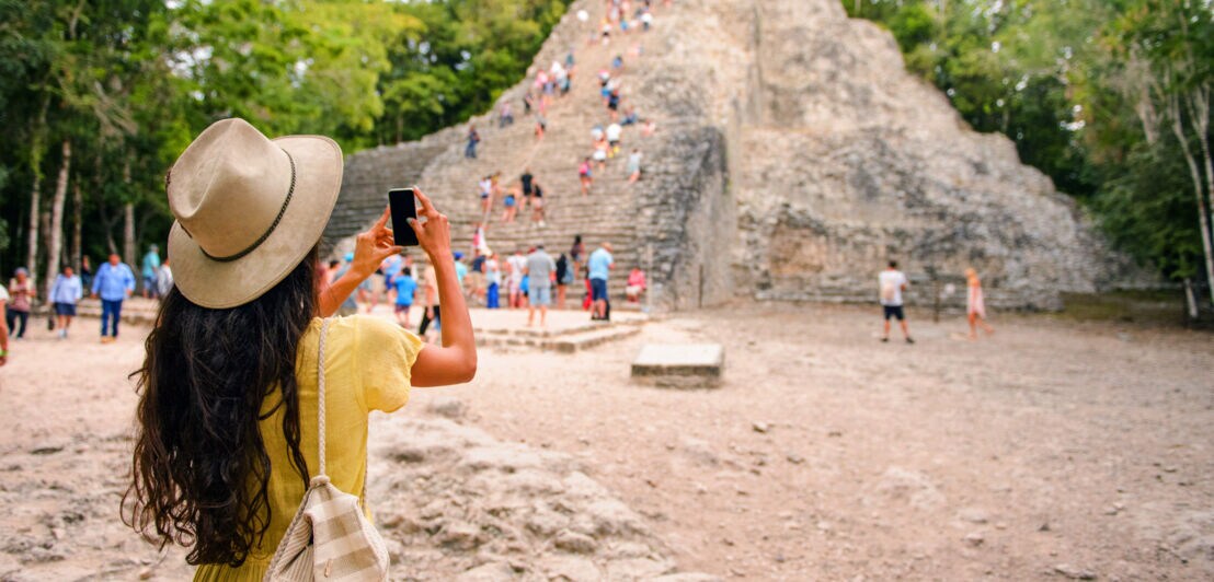 Rückansicht einer Frau mit Strohhut, die mit ihrem Smartphone eine von Personen erklommene Mayapyramide fotografiert